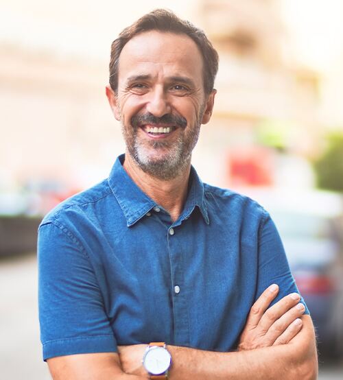 Smiling man in blue shirt with arms crossed outdoors in daylight on a city street.