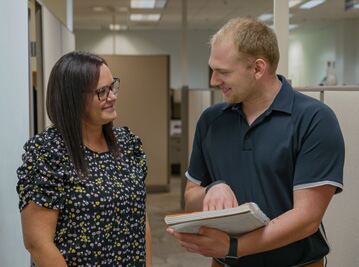 Colleagues review office paperwork and discuss documents in a bright hallway with fluorescent lights.