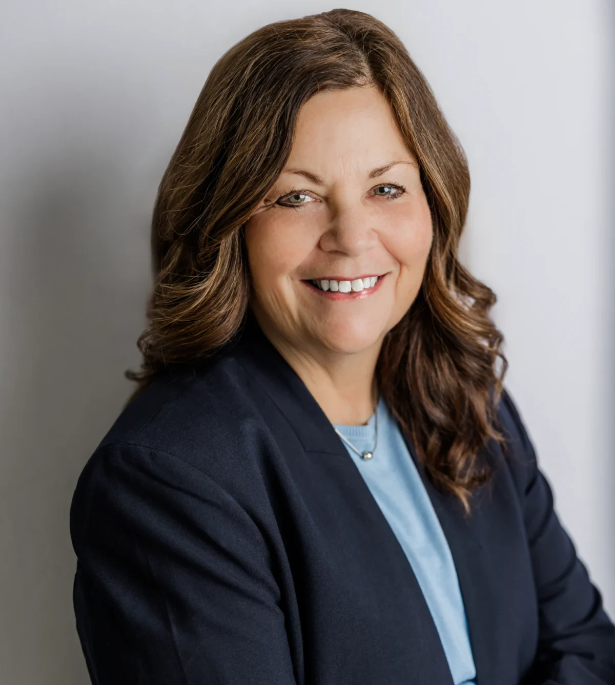 Smiling professional woman in navy blazer and blue blouse, studio headshot on gray background.